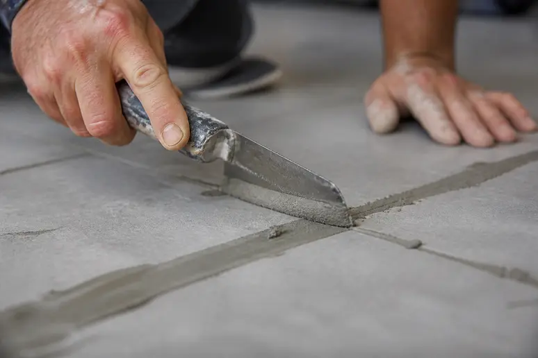 Mains appliquant de l'enduit pour combler les joints de carrelage avant la pose de béton ciré sur carrelage.