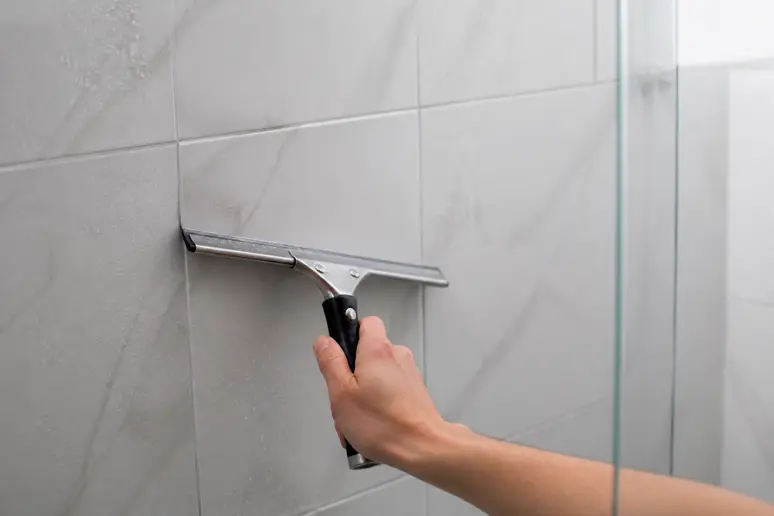 Main utilisant une raclette sur des carreaux de douche pour nettoyer joint carrelage.
